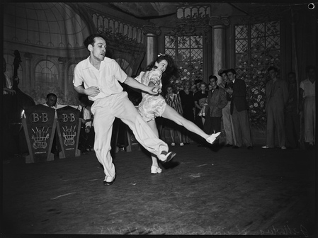 A black-and-white photo of two dancers. They're facing apart, with one leg on the ground, and the other leg pointing up and out (in the same direction). They're connected by a single hand, clearly in the middle of an energetic move.